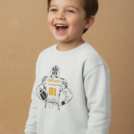 Smiling child wearing a white crewneck sweatshirt with a football graphic on a beige background.