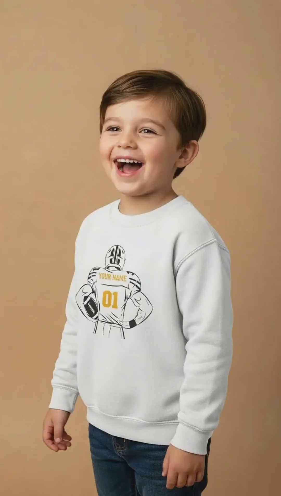 Smiling child wearing a white crewneck sweatshirt with a football graphic on a beige background.